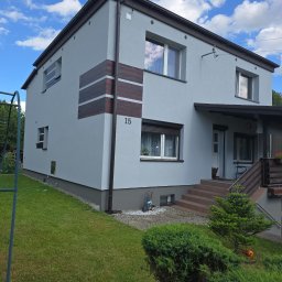 MALBUD - Dwukondygnacyjny dom z szarą elewacją, brązowymi akcentami i numerem 15. Widoczne schody wejściowe, zadbany trawnik i błękitne niebo z chmurami w tle.