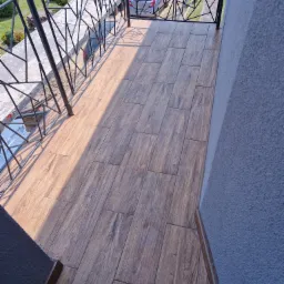 Nowoczesny balkon z drewnopodobną terakotą ułożoną wzdłuż, metalowa balustrada o geometrycznym wzorze, widok na podjazd i zaparkowany samochód.