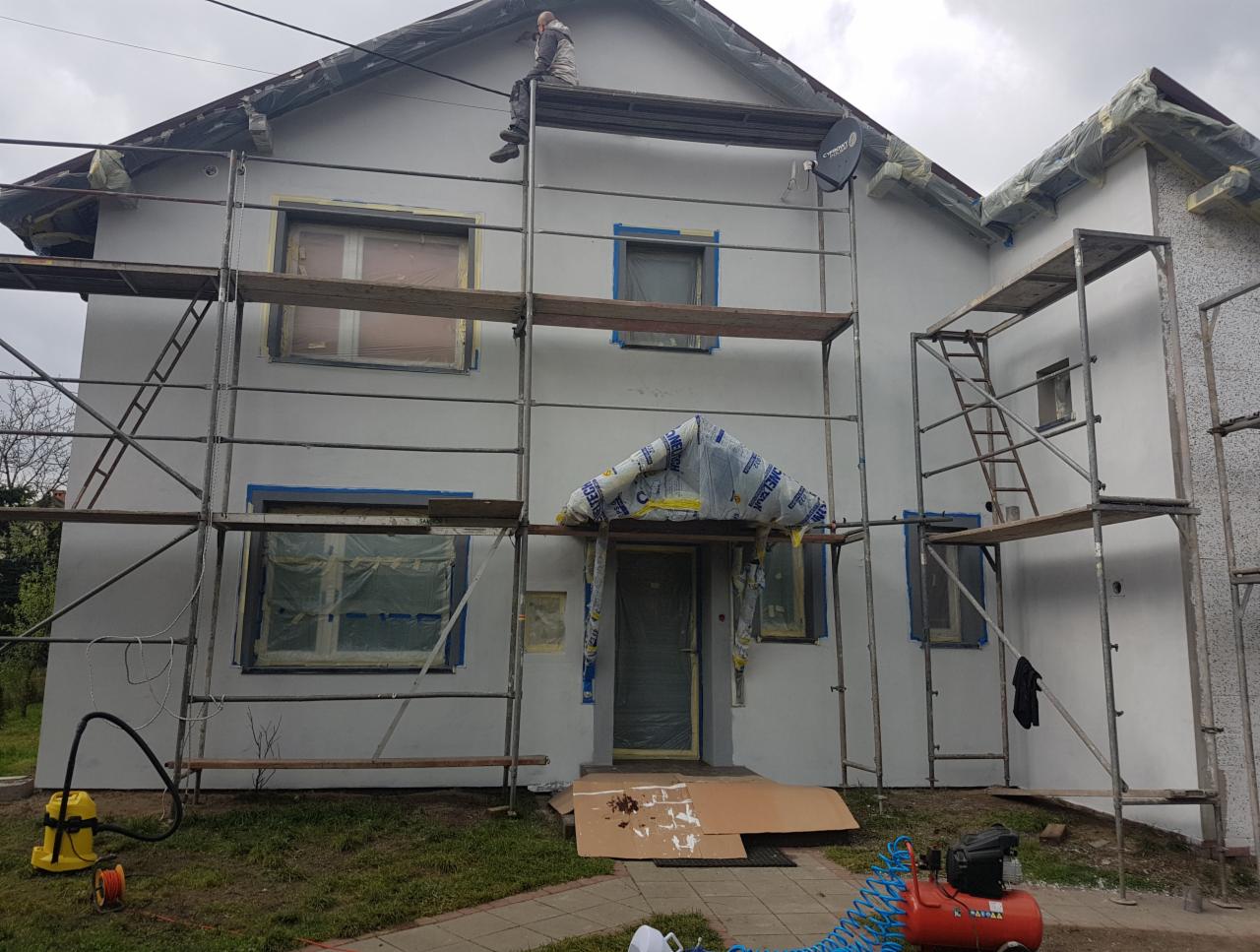 Dwukondygnacyjny dom w trakcie malowania elewacji na szaro, osłonięte okna i drzwi, rusztowania, pracownik siedzący na dachu, kompresor i wąż pneumatyczny na trawniku.