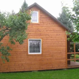 Niewielki, parterowy budynek z drewnianą elewacją w kolorze miodowym, z małym zadaszonym tarasem i dwoma oknami, otoczony zielenią ogrodu.