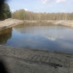 Nowo wykopany staw z wodą, otoczony piaszczystym brzegiem i lasem w tle, z widocznymi śladami opon.