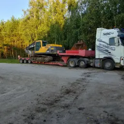Ciężarówka z naczepą niskopodwoziową transportuje żółtą koparkę Volvo w Sępólnie Krajeńskim, widok z boku na tle drzew.