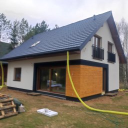M-CONSTRUCTIONS Mariusz Cimochowski - Ocieplony dom jednorodzinny z drewnianą elewacją, widoczne węże do ocieplania, dwukondygnacyjny, z balkonami i grafitową dachówką, w otoczeniu drzew.