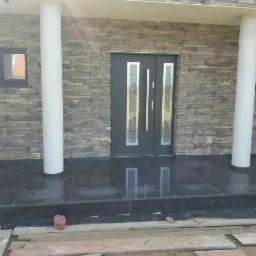 Elewacja domu z kamienia dekoracyjnego, czarne płytki na tarasie, białe kolumny, szare drzwi z przeszkleniami, widoczne deski i kamienie na podłożu.