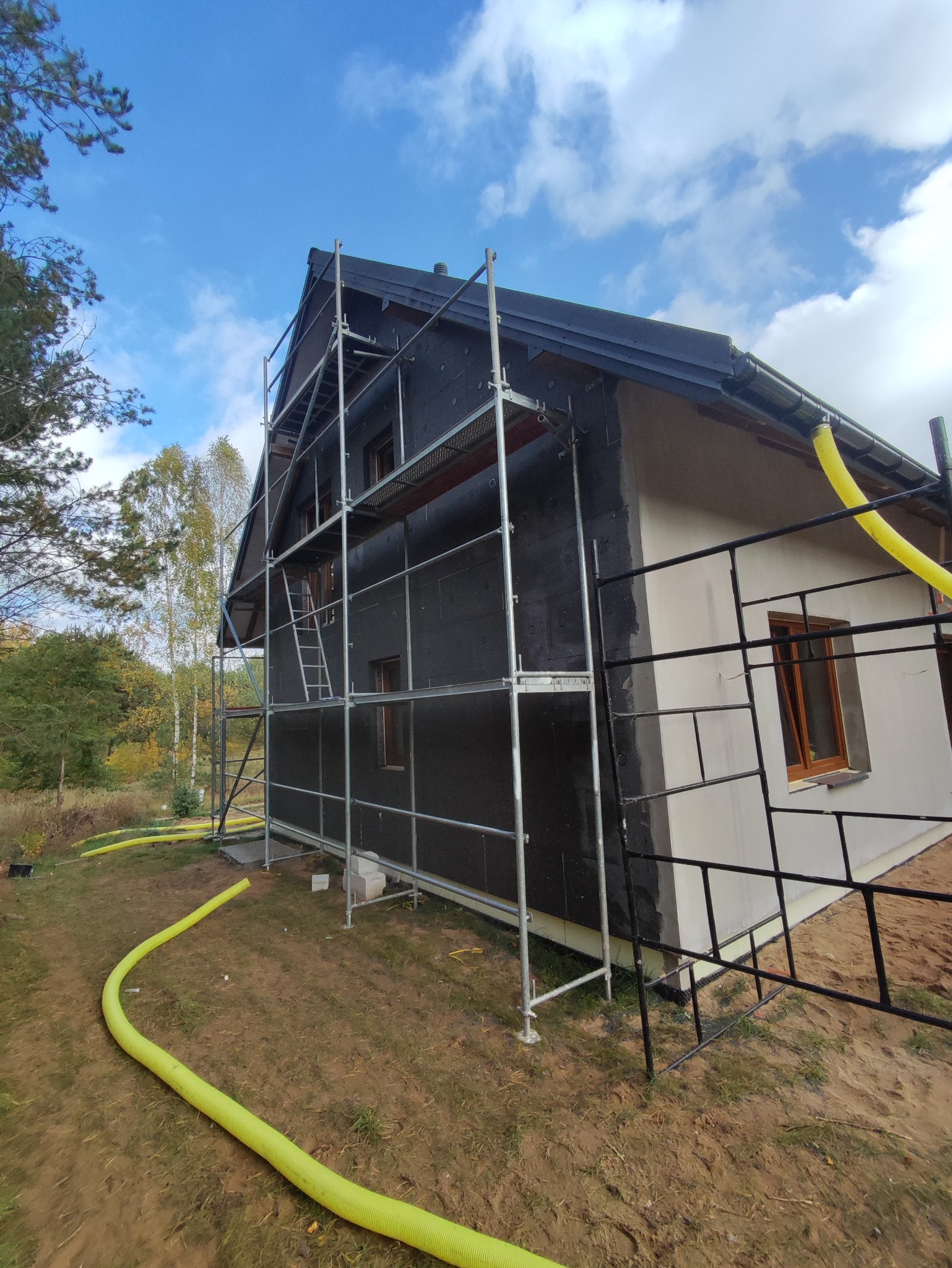 Dom w trakcie ocieplania: czarny styropian na ścianach, rusztowania, fragment otynkowanej elewacji. Żółte węże wentylacyjne na ziemi. Widok z boku, dzień.