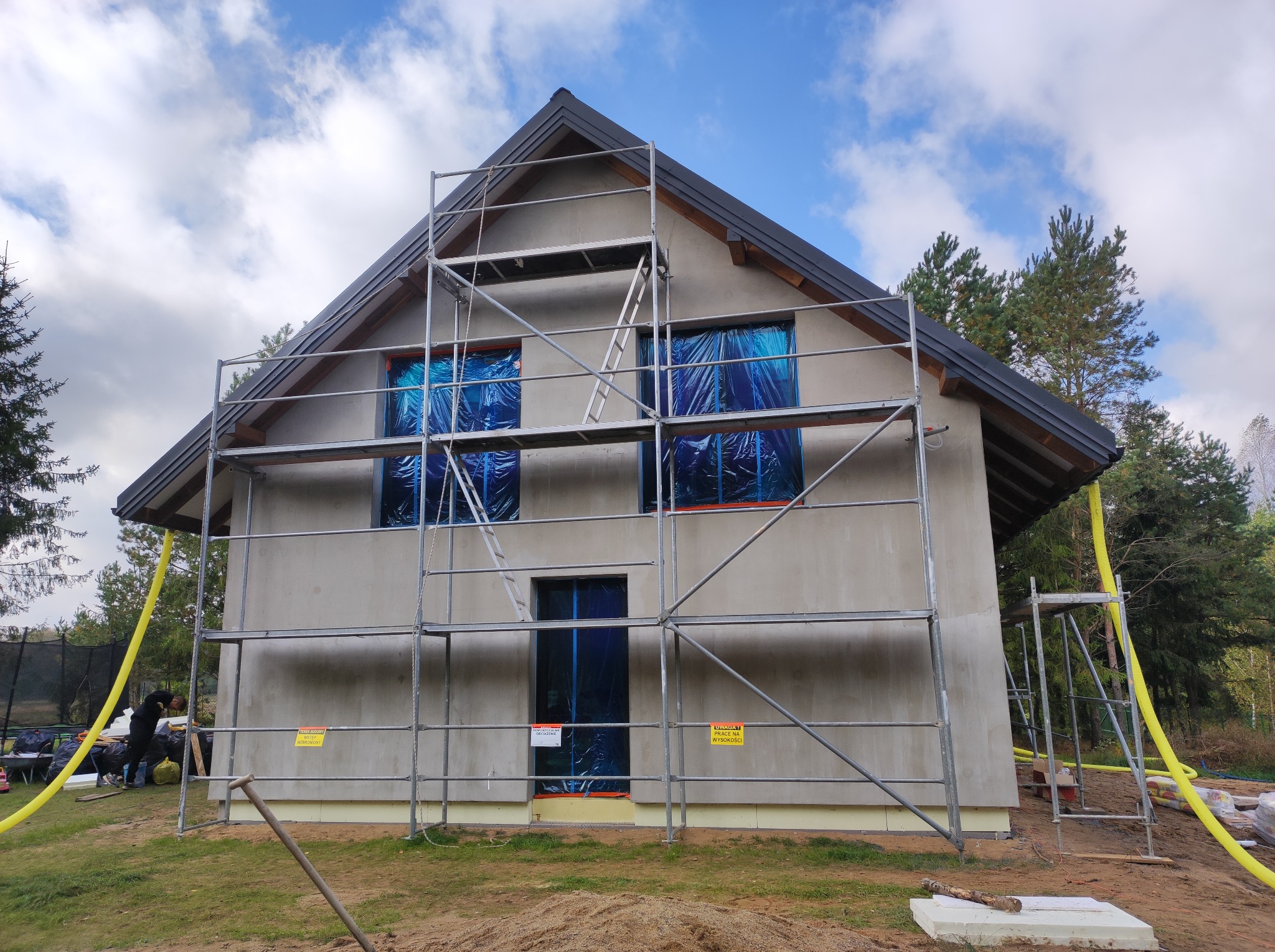 Dom w trakcie ocieplania: szare ściany, rusztowania, okna zabezpieczone folią, widoczne materiały izolacyjne. Prace w toku, widok zewnętrzny, dzień.