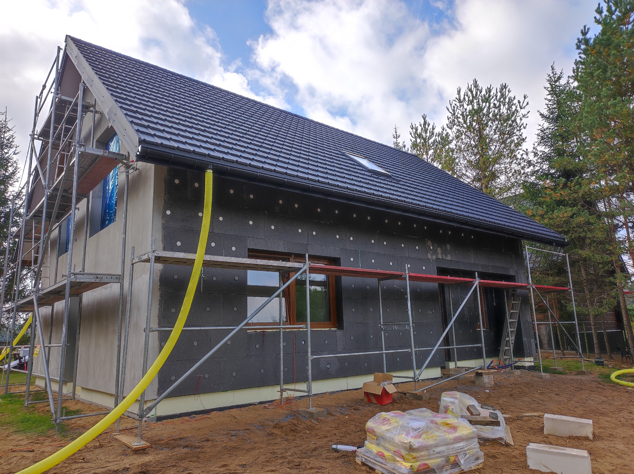 Dom w trakcie ocieplania, widoczne rusztowania i czarny styropian na elewacji, dach pokryty ciemną dachówką. Wokół drzewa i worki z materiałami budowlanymi.