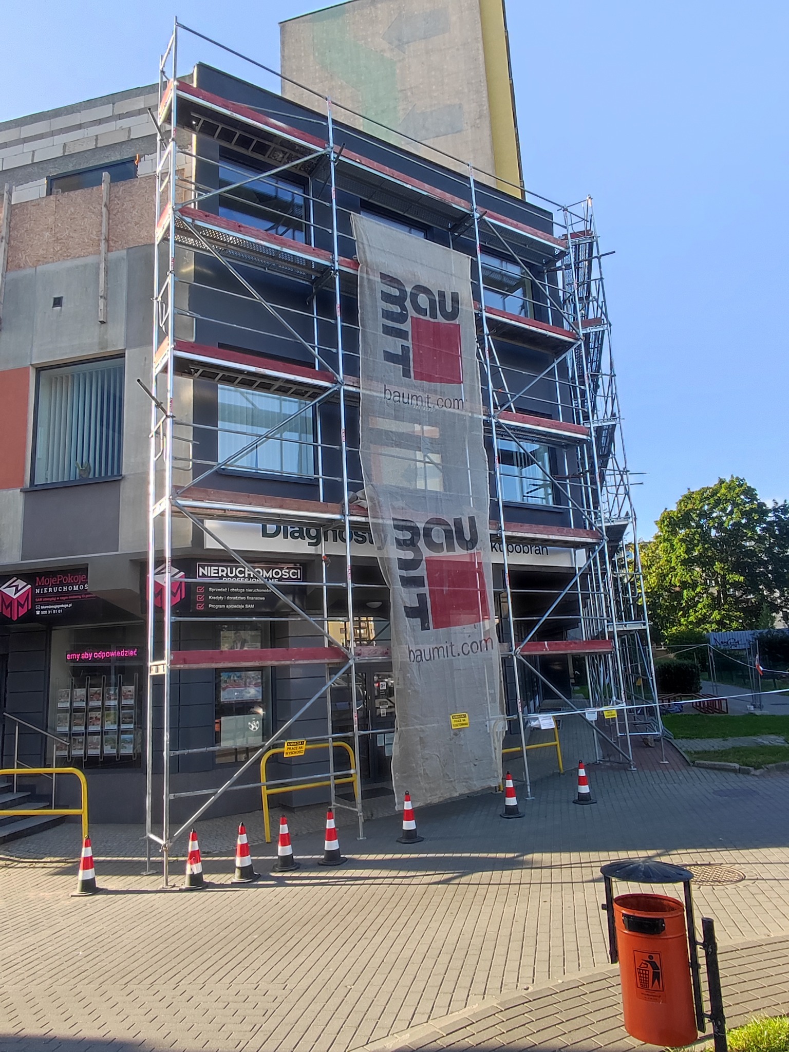 Budynek w rusztowaniach z siatką ochronną Baumit, widoczne okna i fragmenty elewacji. U dołu chodnik z pomarańczowym koszem na śmieci i słupkami ostrzegawczymi.