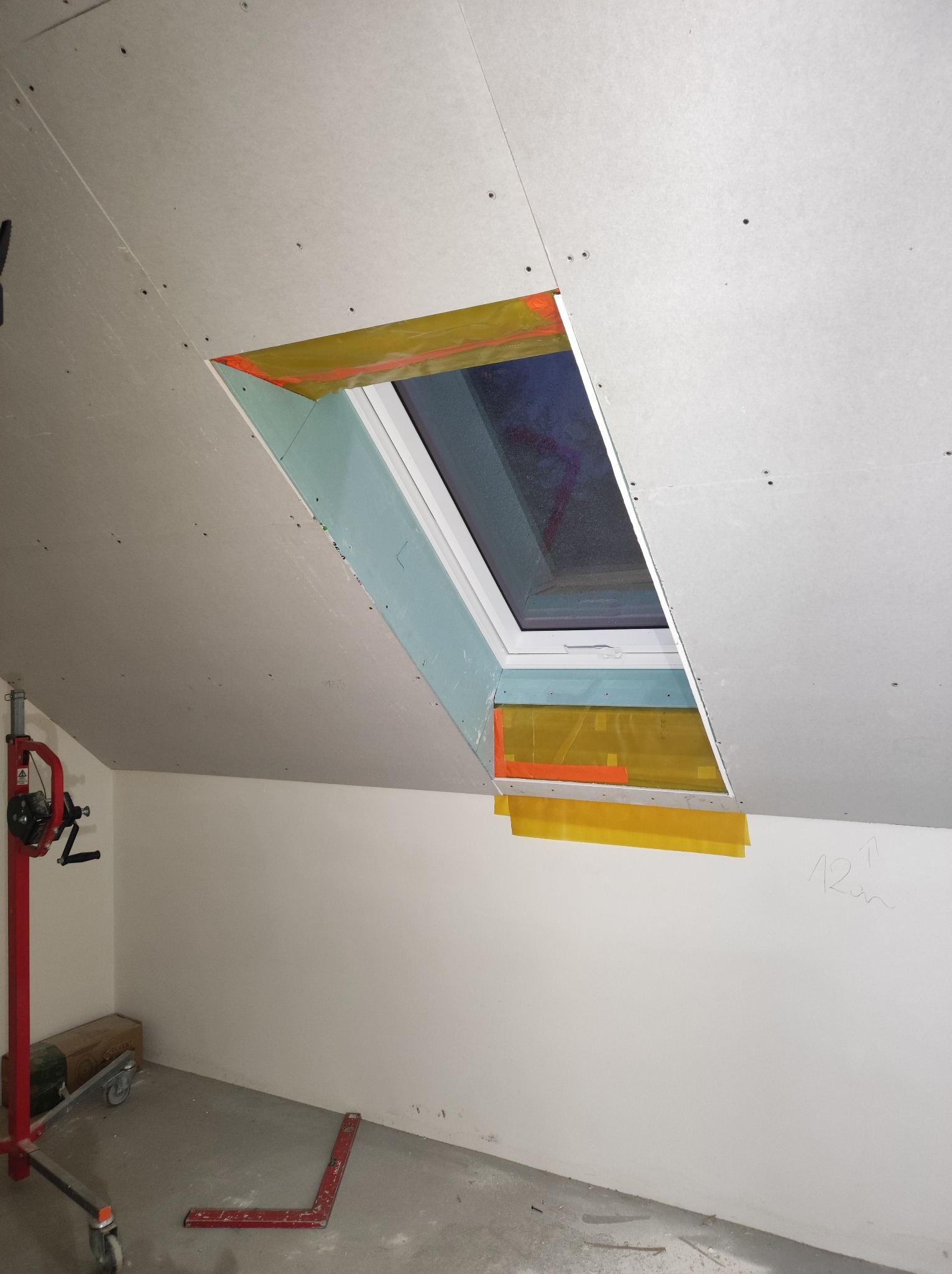 Montaż okna dachowego w płycie kartonowo-gipsowej na poddaszu. Widoczne zabezpieczenia folią i narzędzia. Etap prac przed szpachlowaniem i wykończeniem.