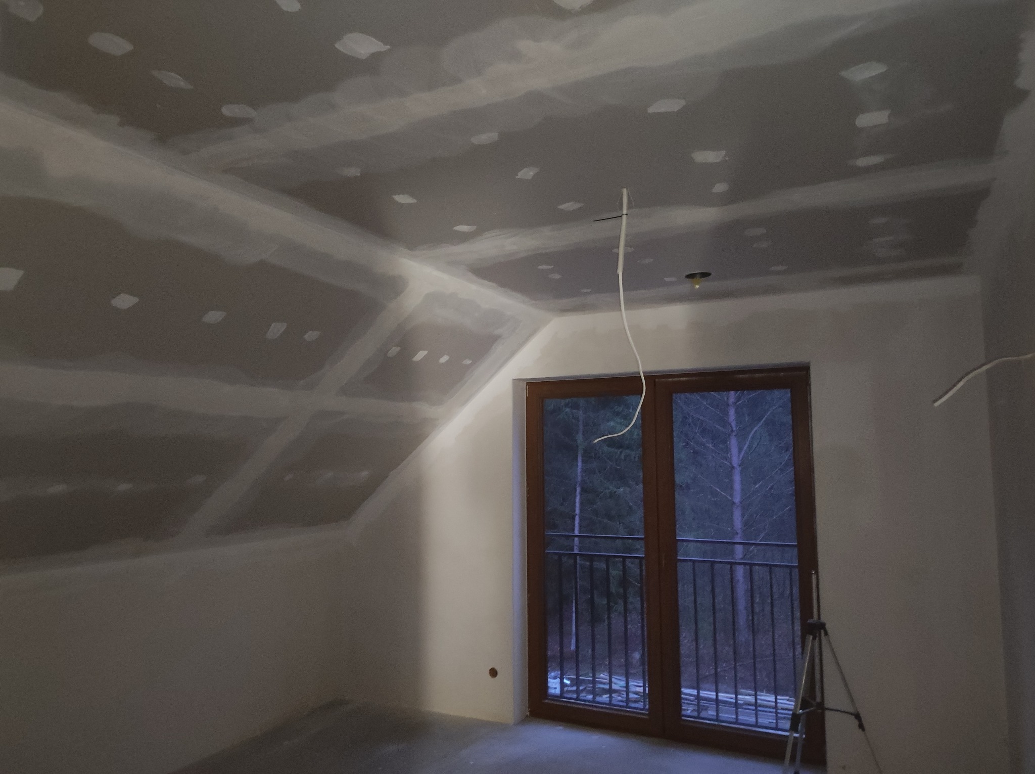 Poddasze po nałożeniu gładzi szpachlowej, widoczne łączenia płyt karton-gips, okno balkonowe z widokiem na las, statyw w rogu pokoju.