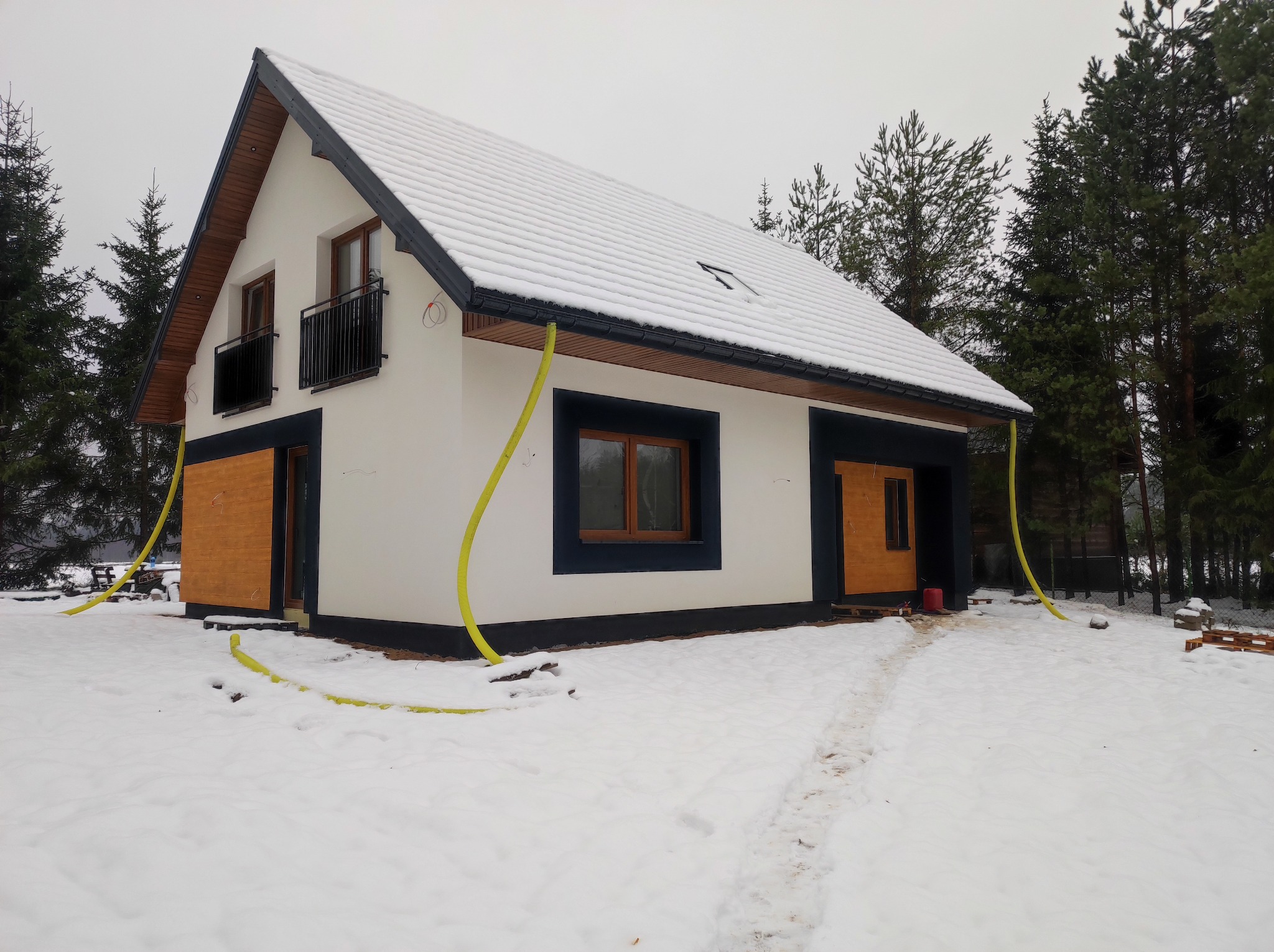 Ocieplony dom jednorodzinny w zimowej scenerii. Biała elewacja kontrastuje z ciemną stolarką okienną i drewnianymi bramami garażowymi. Widoczne węże do ocieplania.