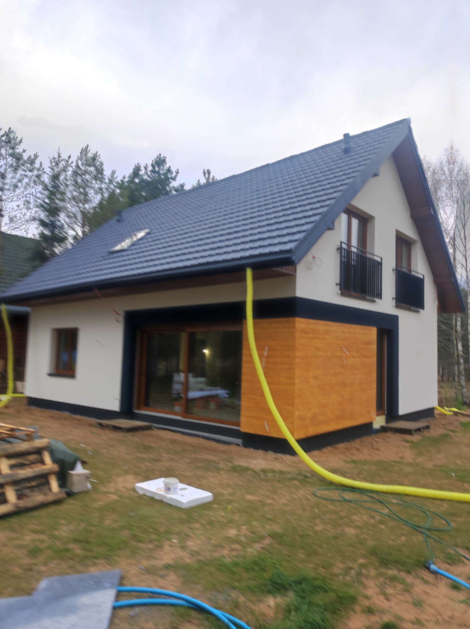 Ocieplony dom jednorodzinny z drewnianą elewacją, widoczne węże do ocieplania, dwukondygnacyjny, z balkonami i grafitową dachówką, w otoczeniu drzew.