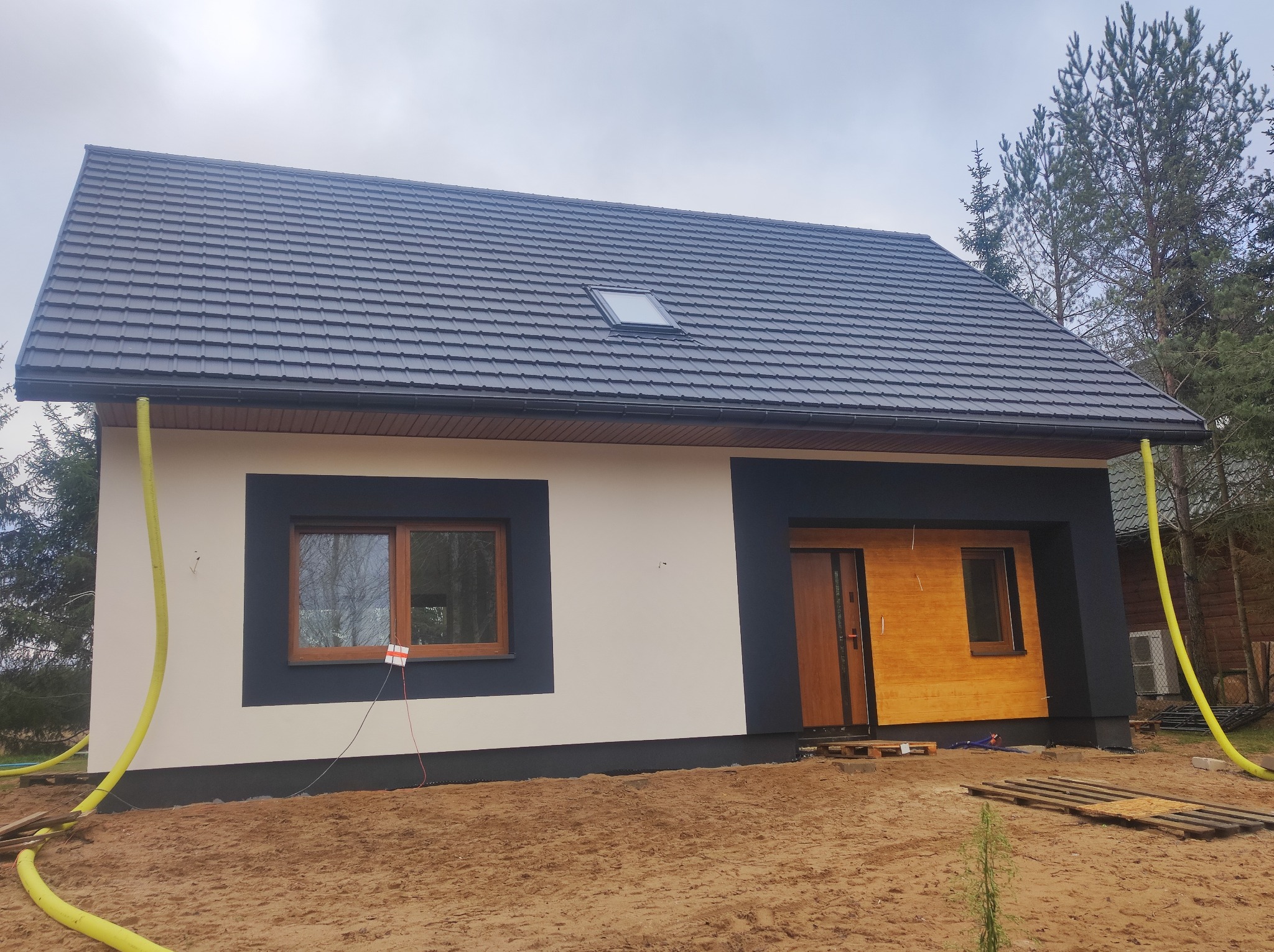 Nowoczesny dom jednorodzinny z ciemną dachówką i jasną elewacją, z widocznymi elementami wykończeniowymi i żółtym wężem na ziemi. Ujęcie z poziomu gruntu.