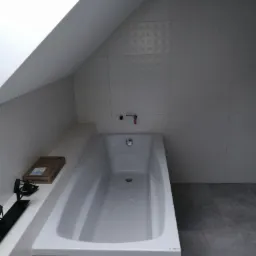Biała, prostokątna wanna obudowana szarymi płytkami w łazience na poddaszu, widoczna armatura i częściowo pudełko po produkcie Geberit, podłoga wyłożona szarymi płytkami.