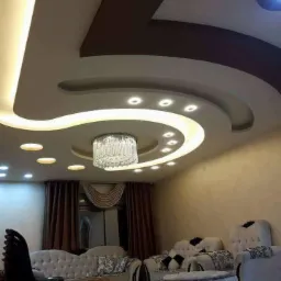 Sufit w salonie z dekoracyjnym podwieszanym elementem, oświetleniem LED i kryształowym żyrandolem, ściany w odcieniach beżu, widoczne meble wypoczynkowe.