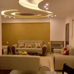 Salon z kremowymi sofami z wzorzystymi poduszkami, oświetleniem punktowym w podwieszanym suficie z gładzią i ścianą w odcieniu musztardowym.