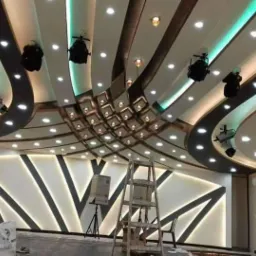Skomplikowany sufit podwieszany z oświetleniem punktowym i dekoracyjnymi pasami, częściowo zakryty drabiną i głośnikiem na statywie, widoczna ściana z geometrycznym wzorem podświetlonym LED.