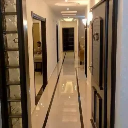 Wykończony korytarz z błyszczącymi, kremowymi płytkami podłogowymi z czarnymi paskami, ciemne futryny drzwiowe i szklane panele po lewej stronie, oświetlenie sufitowe wpuszczone w dekoracyjny sufit.