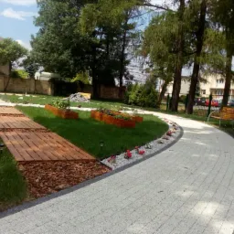 Ogród z drewnianą ścieżką na ściółce, szarą ścieżką z obrzeżem, rabatami kwiatowymi i drewnianymi donicami na trawniku, w tle budynki i drzewa.