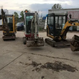 Trzy mini koparki różnych marek zaparkowane na betonowym placu; pierwsza żółta, druga biało-czerwona, trzecia żółto-czarna; w tle białe ciężarówki i budynki magazynowe pod pochmurnym niebem.