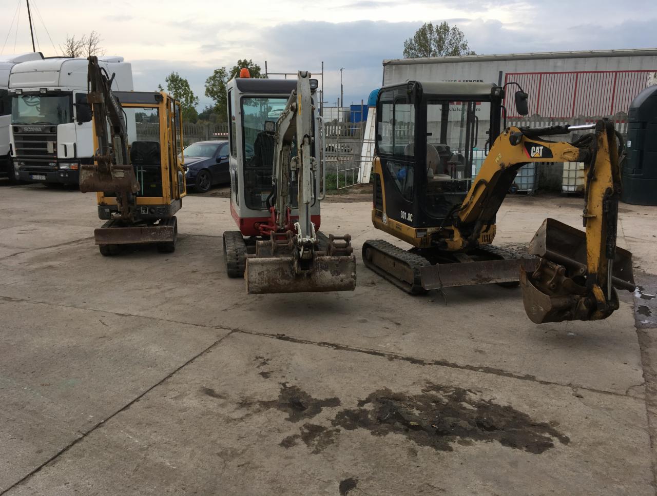Trzy mini koparki różnych marek zaparkowane na betonowym placu; pierwsza żółta, druga biało-czerwona, trzecia żółto-czarna; w tle białe ciężarówki i budynki magazynowe pod pochmurnym niebem.