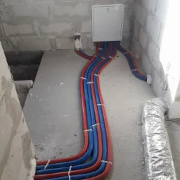 Świeżo ułożone, czerwono-niebieskie rury instalacyjne, zabezpieczone białymi opaskami, wychodzące ze skrzynki w surowym wnętrzu budynku w stanie deweloperskim.
