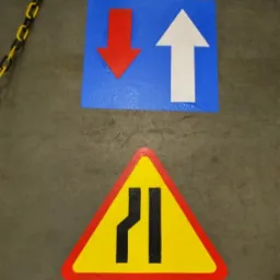 Oznakowanie poziome na szarej posadzce: strzałki kierunkowe (czerwona w dół, biała w górę) na niebieskim kwadracie oraz trójkątny znak ostrzegawczy (zwężenie drogi) w kolorach żółto-czarnym,...