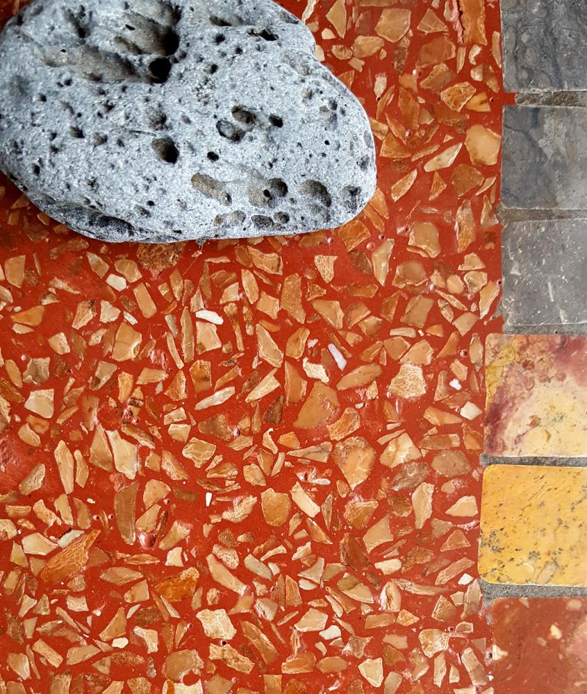 Czerwona posadzka terazzo z wtopionymi kawałkami kamienia w odcieniach beżu i brązu, obok fragmentu posadzki z kolorowych płytek oraz porowaty, szary kamień dekoracyjny.