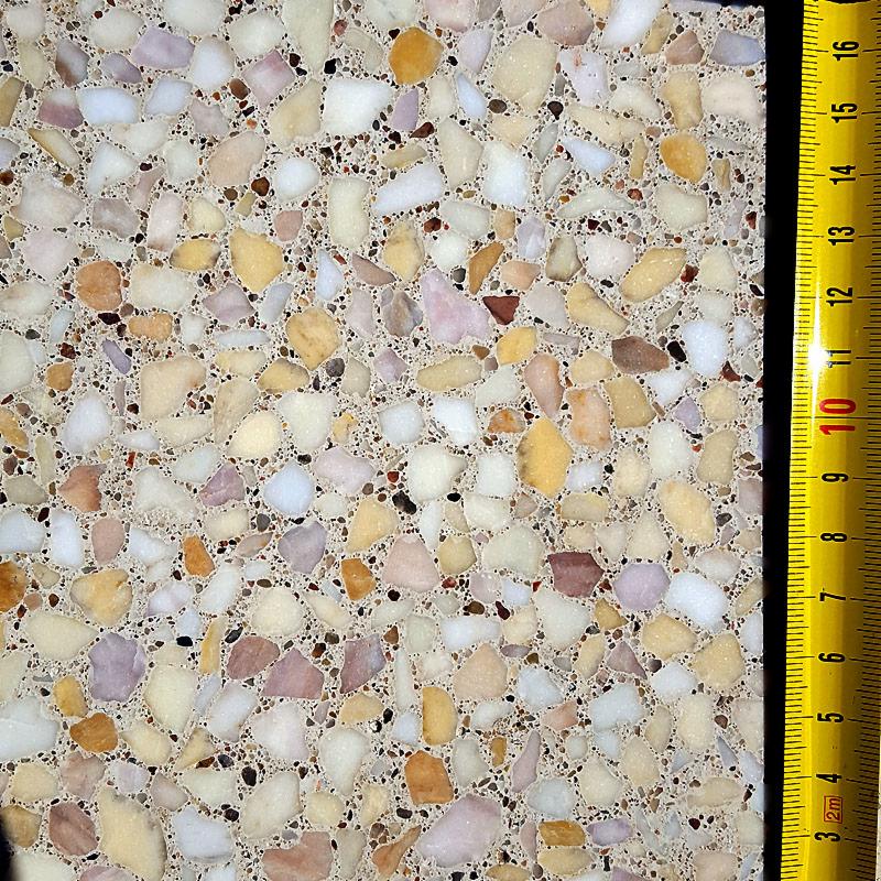 Przekrój próbki posadzki z drobnymi, nieregularnymi kamyczkami w odcieniach bieli, beżu, różu i brązu, z żółtą miarką przy prawej krawędzi.
