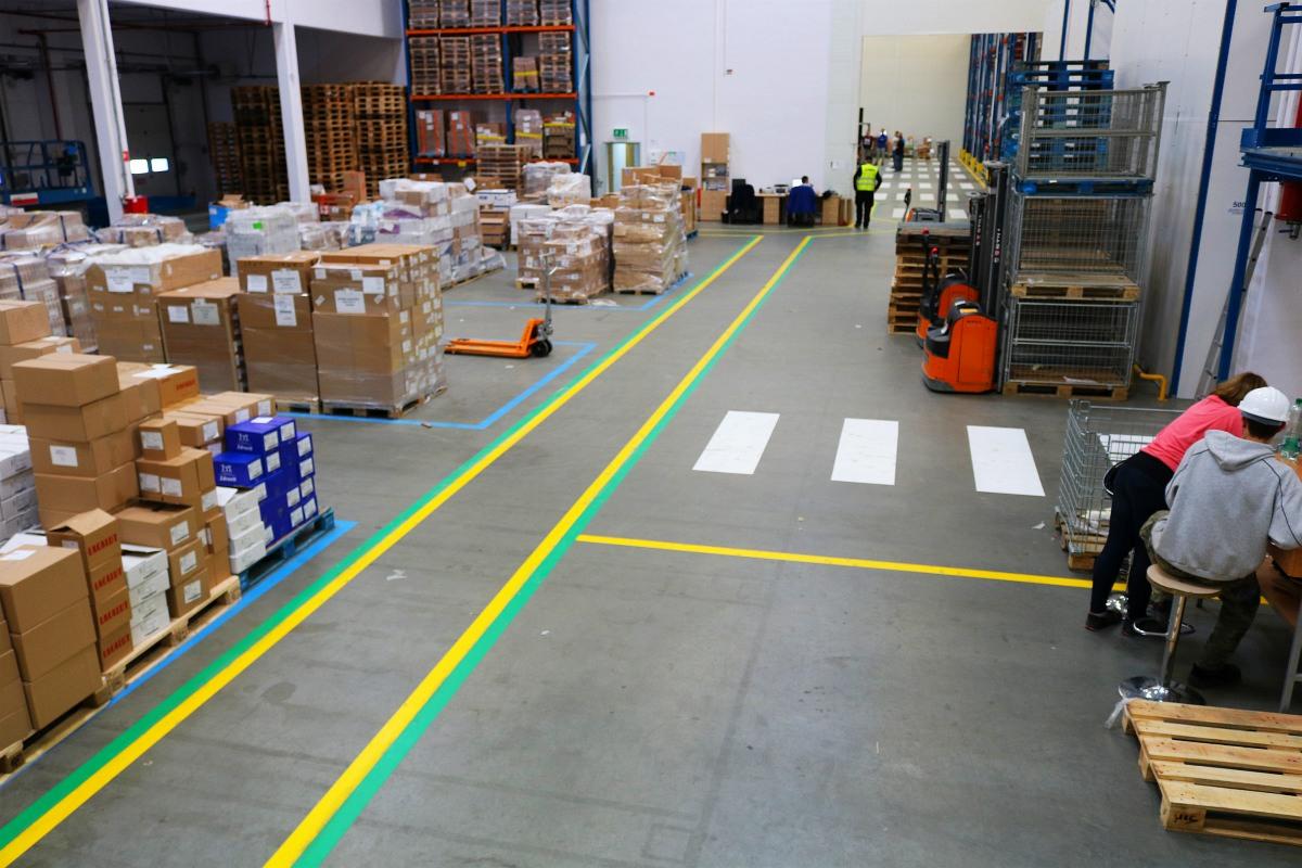 Magazyn z oznakowaniem poziomym: żółto-zielone linie wyznaczają ścieżki, białe pasy przejścia dla pieszych, a niebieskie obrysy miejsca składowania palet z kartonami.