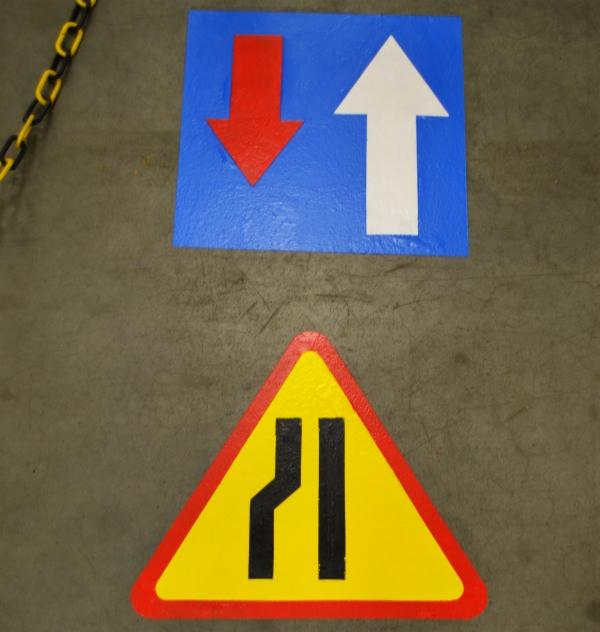 Oznakowanie poziome na szarej posadzce: strzałki kierunkowe (czerwona w dół, biała w górę) na niebieskim kwadracie oraz trójkątny znak ostrzegawczy (zwężenie drogi) w kolorach żółto-czarnym,...