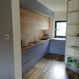 Nowoczesna kuchnia z szarymi szafkami, drewnianym blatem i panelami imitującymi drewno na ścianie, widok na okno i półki z napojami.