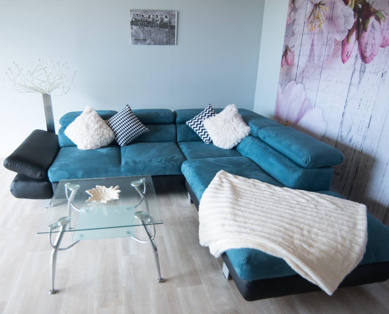 Narożnik z turkusową tapicerką, czarnymi bokami i puszystymi poduszkami w salonie z jasną podłogą i szklanym stolikiem kawowym. Na ścianie widoczny obraz z motywem kwiatowym.