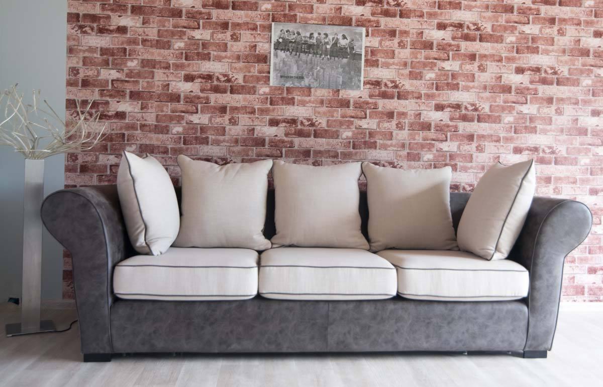 Szara sofa z czterema beżowymi poduszkami na tle ściany z czerwonej cegły i srebrnej dekoracji.