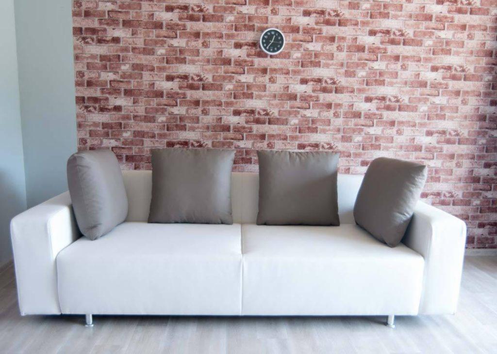 Biała sofa z szarymi poduszkami na tle ściany z czerwonej cegły z zegarem.