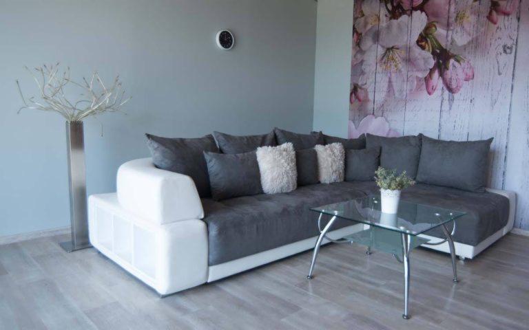 Szary narożnik z białymi bokami i puszystymi poduszkami w salonie z tapetą w kwiaty wiśni i szklanym stolikiem kawowym.