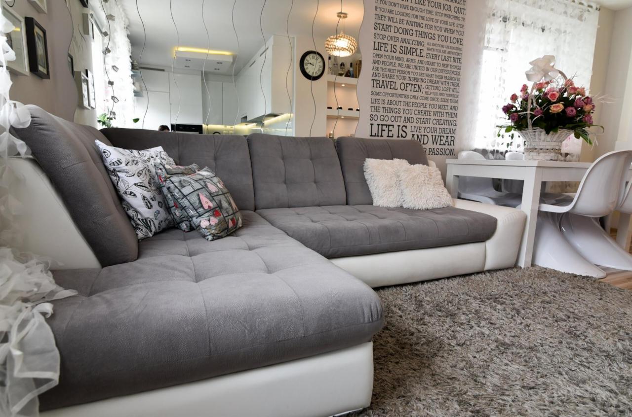 Szary narożnik tapicerowany z białymi bokami, dekoracyjne poduszki, puszysty dywan i biały stół z kwiatami w słonecznym wnętrzu.