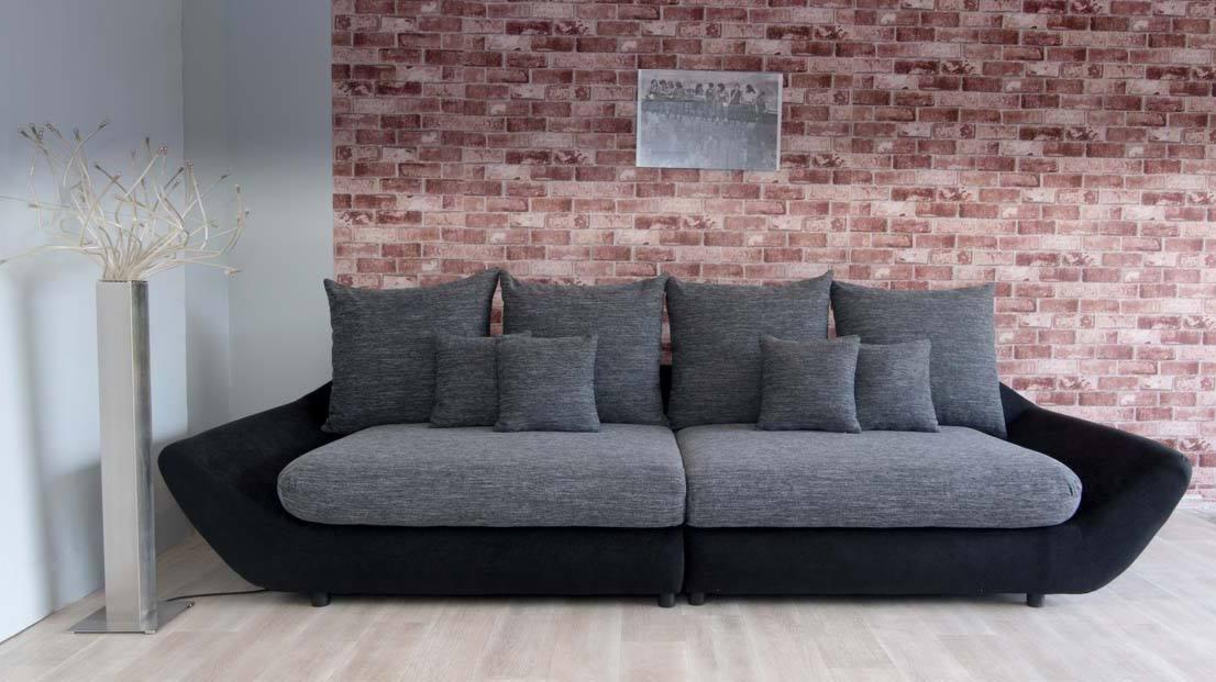 Nowoczesna sofa modułowa w odcieniach szarości i czerni, z siedmioma poduszkami, ustawiona na tle ściany z czerwonej cegły i srebrnego wazonu z dekoracyjnymi gałązkami.