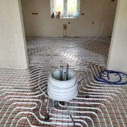 Białe rury ogrzewania podłogowego rozłożone na czerwonej siatce podłogowej w surowym wnętrzu domu, zw&oacute;j rur na metalowym stojaku w pierwszym planie, okno w tle.