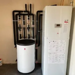 Biały, nowoczesny piec gazowy dwufunkcyjny z zasobnikiem wody, połączony czarnymi rurami instalacji grzewczej, na tle beżowej ściany i ciemnej podłogi z płytek.