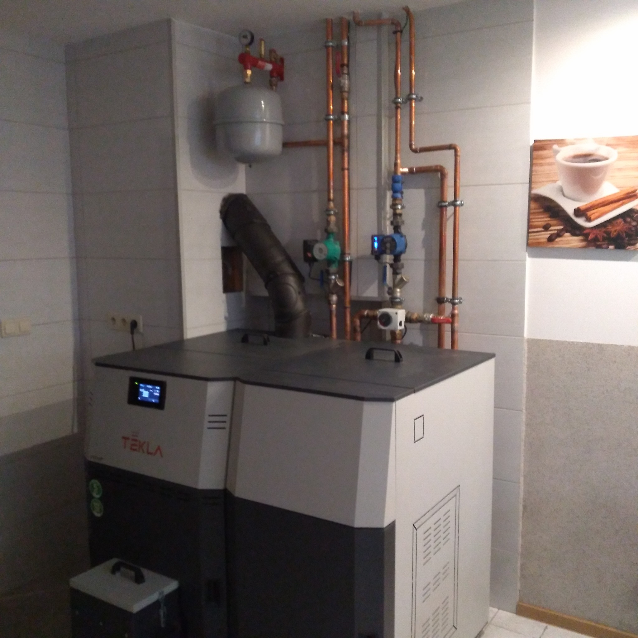 Nowoczesny piec grzewczy Tekla z podajnikiem, z widoczną instalacją hydrauliczną i zbiornikiem wyrównawczym, umieszczony w pomieszczeniu z wykończeniem ceramicznym.