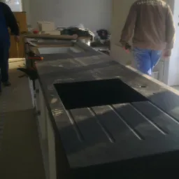 Czarny blat kuchenny z wyciętym otworem na zlew i ociekaczem, w trakcie montażu, z widocznymi narzędziami monterskimi i pracownikami w tle.