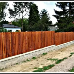 Nowoczesne ogrodzenie drewniane z podmurówką z kamienia łupanego na tle zieleni. Słupki ogrodzeniowe z szarego kamienia. Widok na posesję za ogrodzeniem.