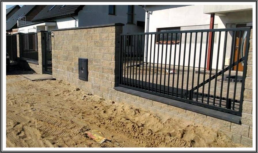 Fragment ogrodzenia z metalowymi prętami i murkiem z beżowych bloczków, skrzynka pocztowa na murku, teren wokół wyrównany piaskiem, w tle budynek mieszkalny.