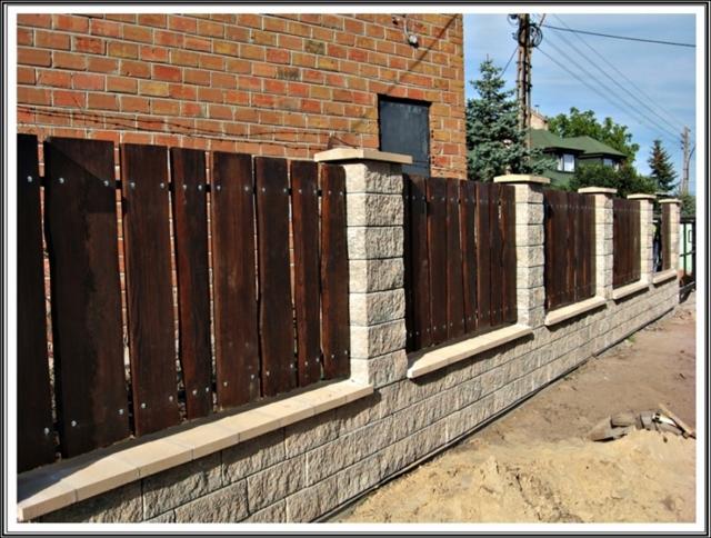 Nowoczesne ogrodzenie z drewnianymi panelami i kamiennymi słupkami. Widok na ogrodzenie wzdłuż posesji, z ceglanym budynkiem w tle i zielenią w oddali.