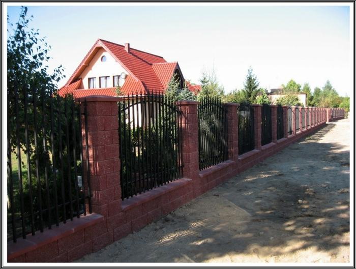 Długie, czarne metalowe ogrodzenie z ceglanymi słupkami i podmurówką, chroniące posesję z domem o czerwonym dachu w tle.