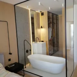 Nowoczesna łazienka z wolnostojącą białą wanną, czarną armaturą i szklaną ścianą oddzielającą ją od garderoby z przesuwnymi drzwiami.