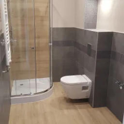 Nowoczesna łazienka z kabiną prysznicową z drewnianymi panelami, podwieszaną toaletą i szarymi płytkami z dekoracyjnym pasem, oświetlona punktowo.