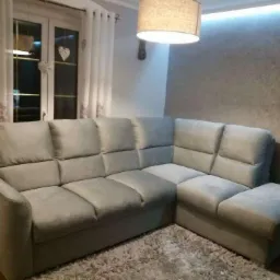 Szary narożnik z poduszkami w salonie, na dywanie shaggy. Lampa sufitowa, okno z firankami i serduszkiem. Neutralna kolorystyka wnętrza.