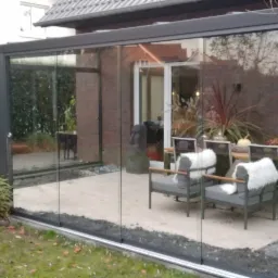 Zadaszony taras z przesuwnymi szklanymi ścianami, wyposażony w meble ogrodowe i dekoracje, widok z zewnątrz na ogród z zielenią i parasolem.